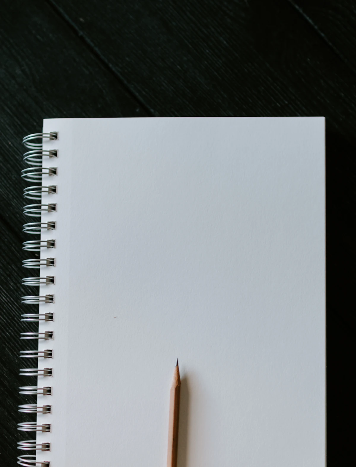 A blank spiral notebook and a sharpened pencil on a dark wooden table