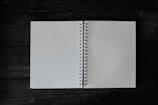 An open spiral notebook with blank white pages rests on a dark wooden surface. The notebook is centrally positioned, creating a contrast between the light pages and the dark, textured background.