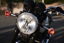A close-up view of a motorcycle headlight with two red front indicators. The headlight is round and metallic, reflecting the surrounding environment. The handlebars are visible with black rubber grips, set against a blurred background.