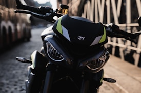 A close-up of a modern motorcycle with a sleek design is parked on a cobblestone street. The vehicle has a black and neon green color scheme with prominent headlights and the letters 'RS' on the front. The background shows a blurred view of buildings and a bridge railing, suggesting an urban setting.