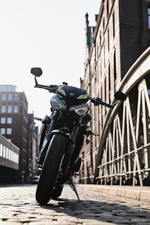 A motorcycle is parked on a cobblestone street with a backdrop of tall, brick buildings and an iron bridge. The sunlight creates a strong contrast, emphasizing the contours of the motorcycle with clear shadows and highlights.