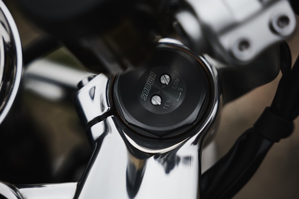 The image features a close-up of a metallic mechanical component, likely part of a motorcycle or bicycle, with a focus on a dial labeled 'SHOWA'. The dial includes markings for adjustments labeled 'TEN', 'COM', and 'H S'. The shiny, reflective surfaces suggest a polished metal material.