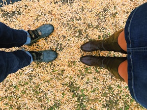 Two pairs of feet are standing on a ground covered with scattered corn kernels. The footwear includes one pair of black boots and another pair of green boots.