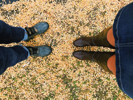 Two pairs of feet are standing on a ground covered with scattered corn kernels. The footwear includes one pair of black boots and another pair of green boots.