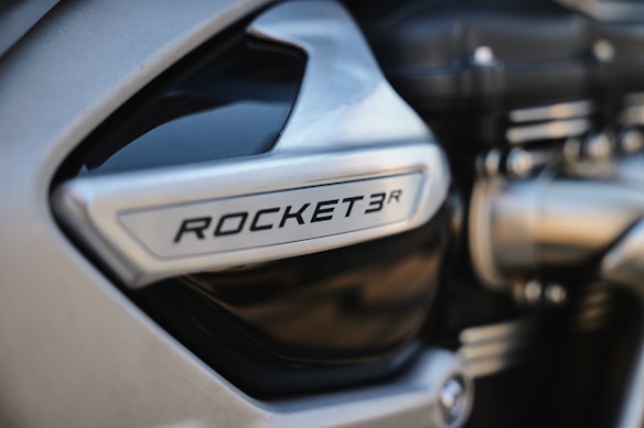 Close-up of a motorcycle engine with the text 'ROCKET 3R' on a metallic surface, showcasing intricate mechanical parts and a smooth, modern design.