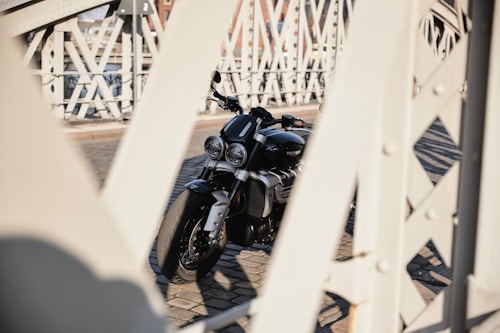 A motorcycle is parked on a cobblestone surface, framed by the metal structure of a bridge. The motorcycle is black and chrome, with two large front headlights and a muscular design. The bridge features diagonal and vertical metal beams, and the background includes parts of urban buildings.