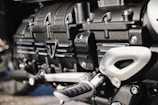 Close-up view of a metallic engine component, featuring intricately designed parts with bolts, grooves, and a triangular emblem. The metallic surfaces have a polished finish, reflecting light subtly.
