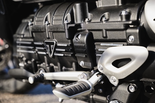 Close-up view of a metallic engine component, featuring intricately designed parts with bolts, grooves, and a triangular emblem. The metallic surfaces have a polished finish, reflecting light subtly.