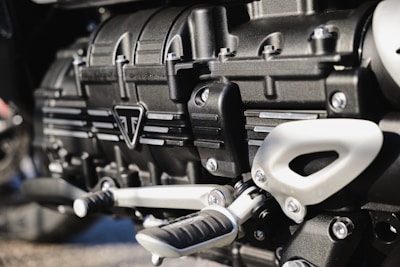 Close-up view of a metallic engine component, featuring intricately designed parts with bolts, grooves, and a triangular emblem. The metallic surfaces have a polished finish, reflecting light subtly.