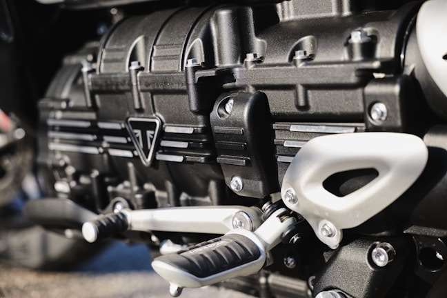 Close-up view of a metallic engine component, featuring intricately designed parts with bolts, grooves, and a triangular emblem. The metallic surfaces have a polished finish, reflecting light subtly.
