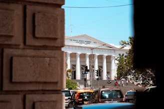 A grand white building with columns and a triangular pediment, identified as 'The Asiatic Society, Mumbai'. The building is seen from between two large stone blocks in the foreground. There are several people walking on the steps in front of the building. A few cars and a traffic light are visible at the bottom of the steps. Trees are partially visible on the right and left sides of the image, and the sky is clear and blue.