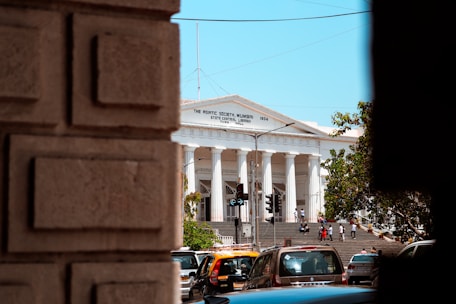 A grand white building with columns and a triangular pediment, identified as 'The Asiatic Society, Mumbai'. The building is seen from between two large stone blocks in the foreground. There are several people walking on the steps in front of the building. A few cars and a traffic light are visible at the bottom of the steps. Trees are partially visible on the right and left sides of the image, and the sky is clear and blue.