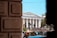 A grand white building with columns and a triangular pediment, identified as 'The Asiatic Society, Mumbai'. The building is seen from between two large stone blocks in the foreground. There are several people walking on the steps in front of the building. A few cars and a traffic light are visible at the bottom of the steps. Trees are partially visible on the right and left sides of the image, and the sky is clear and blue.