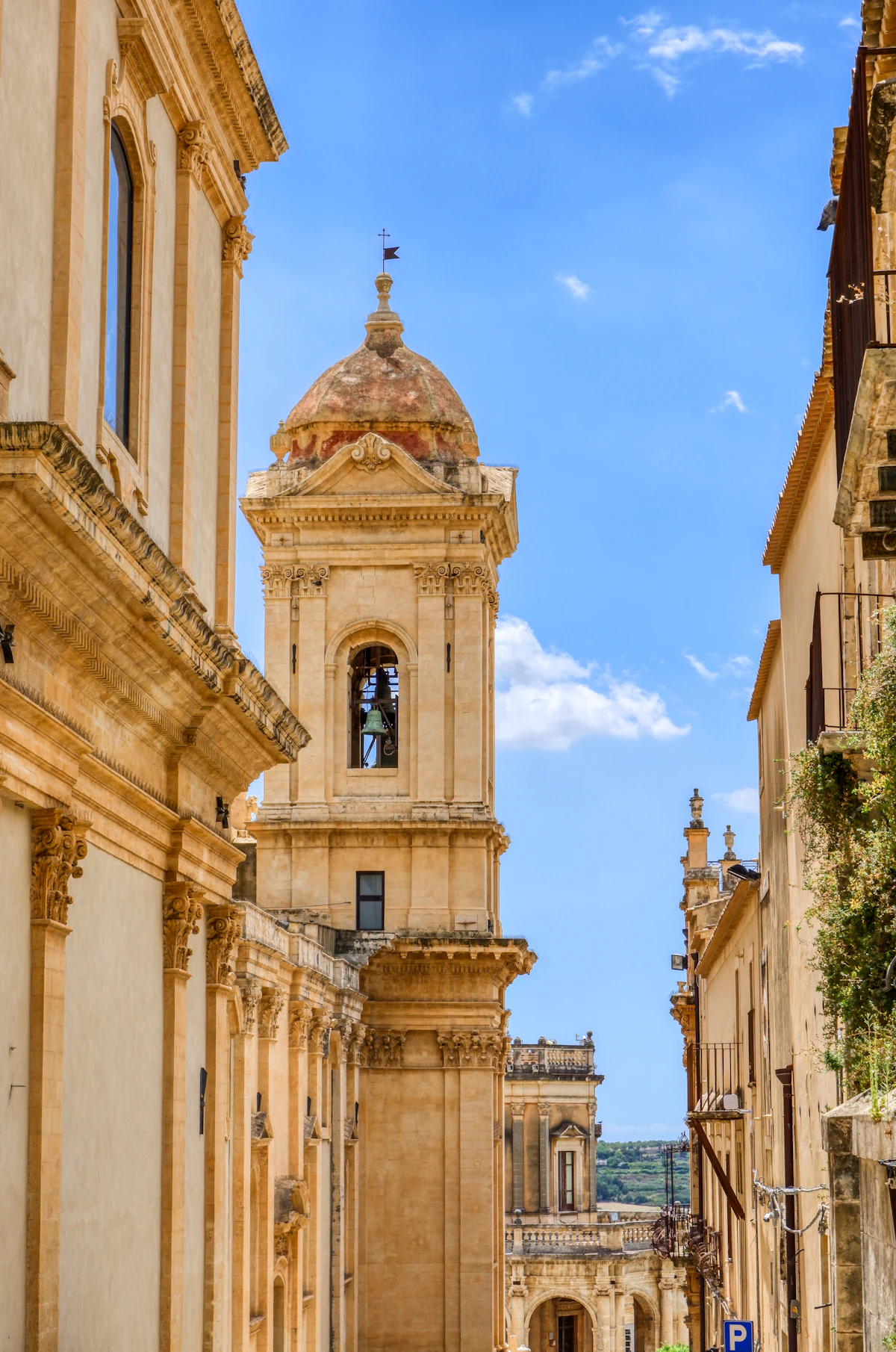 A baroque bell tower rising above a narrow sunlit street in Sicily
