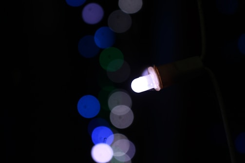 A single white LED bulb is lit against a dark background, surrounded by out-of-focus bokeh lights in various shades of blue and green.