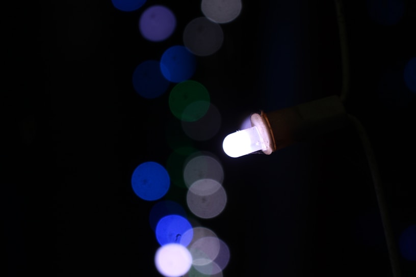 A single white LED bulb is lit against a dark background, surrounded by out-of-focus bokeh lights in various shades of blue and green.