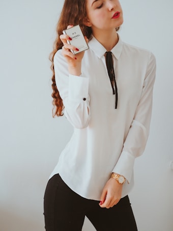 A woman with long, wavy hair holds a bottle of Chanel No. 5 perfume close to her face. She wears a white blouse with a black ribbon tie and a gold watch on her wrist. Her lips are painted red, matching the color of her fingernails.