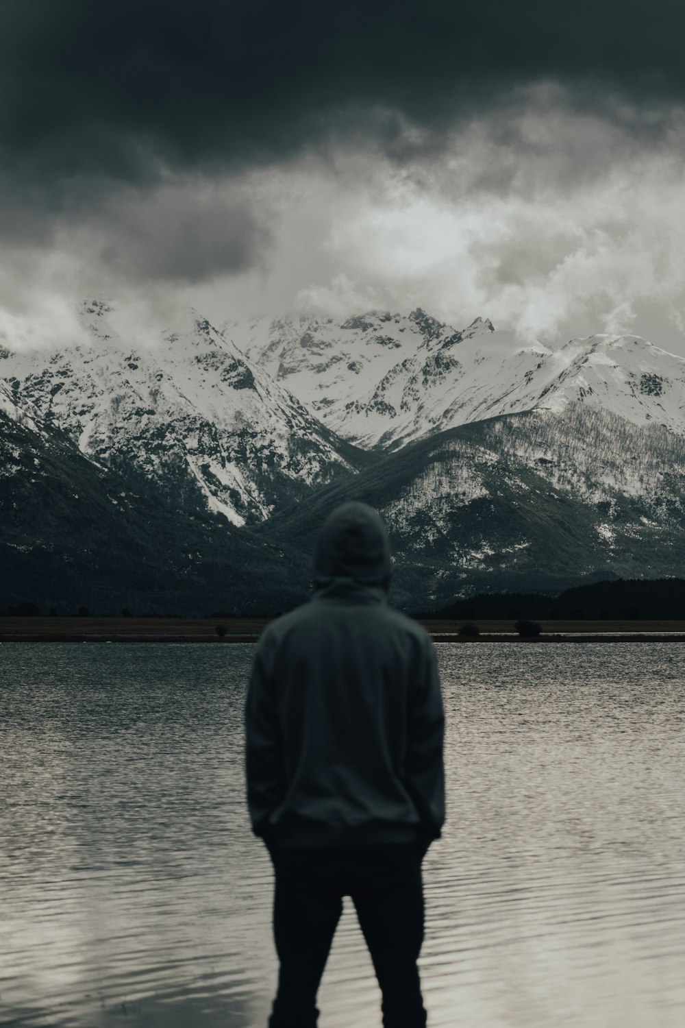 uomo in felpa con cappuccio grigia in piedi vicino allo specchio d'acqua e alla montagna coperta di neve durante il giorno