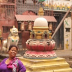 A woman in a colorful outfit is smiling in front of a vibrant, ornate stupa, surrounded by traditional Nepalese architectural elements and a lion statue in a courtyard.