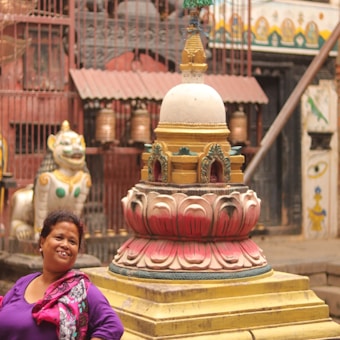 A woman in a colorful outfit is smiling in front of a vibrant, ornate stupa, surrounded by traditional Nepalese architectural elements and a lion statue in a courtyard.