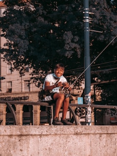 A person sitting on a stone ledge, smiling while holding a fishing rod. The background consists of large, leafy trees and a section of an urban environment, including buildings and street elements such as a utility pole.
