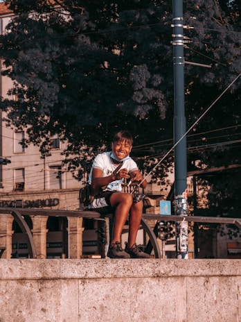 A person sitting on a stone ledge, smiling while holding a fishing rod. The background consists of large, leafy trees and a section of an urban environment, including buildings and street elements such as a utility pole.
