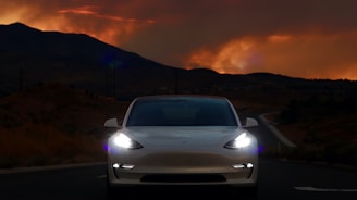 A white car with illuminated headlights is positioned at the center of an empty road, with a dramatic, fiery orange sunset above silhouetted mountains in the background. The scene conveys a sense of solitude and focus on the vehicle.