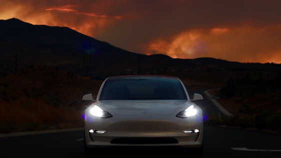 A white car with illuminated headlights is positioned at the center of an empty road, with a dramatic, fiery orange sunset above silhouetted mountains in the background. The scene conveys a sense of solitude and focus on the vehicle.