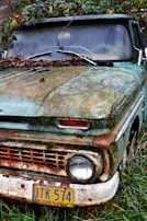 An old, rusty vehicle partially covered in moss and leaves, situated against a backdrop of overgrown vegetation. The license plate indicates it is from Oregon and displays the number JTK 574. The exterior is weathered, showing signs of neglect and exposure to the elements over time.