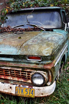 An old, rusty vehicle partially covered in moss and leaves, situated against a backdrop of overgrown vegetation. The license plate indicates it is from Oregon and displays the number JTK 574. The exterior is weathered, showing signs of neglect and exposure to the elements over time.