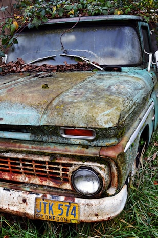An old, rusty vehicle partially covered in moss and leaves, situated against a backdrop of overgrown vegetation. The license plate indicates it is from Oregon and displays the number JTK 574. The exterior is weathered, showing signs of neglect and exposure to the elements over time.