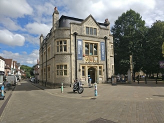 A historic building with stone walls and arched windows stands prominently at the corner of a cobblestone street. Banners hanging on the building indicate it is a museum. Several pedestrians and a person on a motorcycle are visible nearby, with trees providing a lush green backdrop. The sky is partly cloudy, adding to the scene's ambient light.