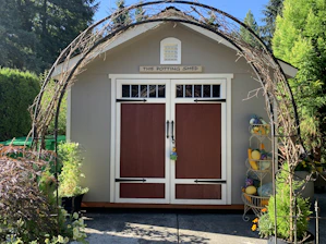 A quaint potting shed with brown double doors is framed by a decorative metal arch entwined with vines. The shed sits on a concrete path, surrounded by lush greenery and plants, including a variety of potted flowers. To the right, there's a tiered stand filled with colorful gourds.