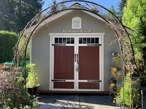 A quaint potting shed with brown double doors is framed by a decorative metal arch entwined with vines. The shed sits on a concrete path, surrounded by lush greenery and plants, including a variety of potted flowers. To the right, there's a tiered stand filled with colorful gourds.