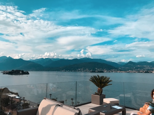 A scenic rooftop terrace overlooks a calm body of water, with an island and mountainous landscape in the background. The sky is partially cloudy and a bright blue, enhancing the tranquil atmosphere. The terrace is equipped with modern outdoor furniture and a potted palm plant.