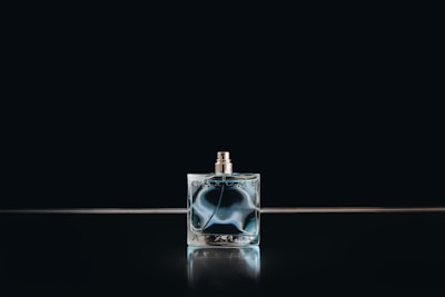 A glass perfume bottle with a metallic cap stands against a dark background. The bottle has an intricate, swirling bluish design within the glass, reflecting light softly.