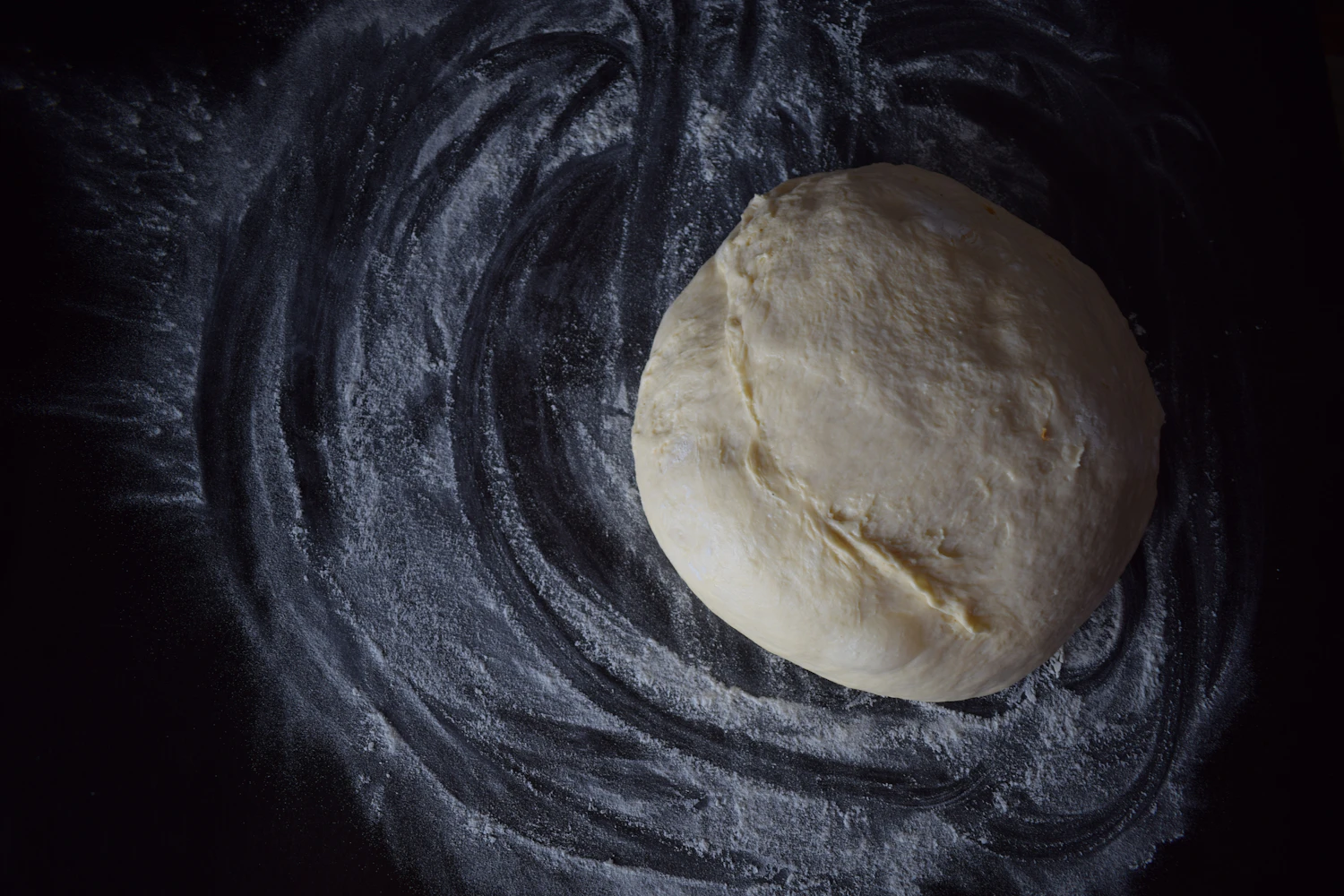 A pale ball of dough resting on a dark worktop swept with flour