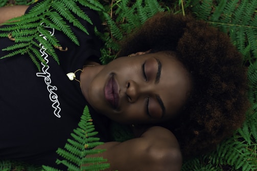 A person with closed eyes rests peacefully among green ferns, wearing a black shirt with the word 'champion' in white lettering. Their head is positioned on their arm, and they have a calm, serene expression.