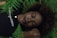 A person with closed eyes rests peacefully among green ferns, wearing a black shirt with the word 'champion' in white lettering. Their head is positioned on their arm, and they have a calm, serene expression.