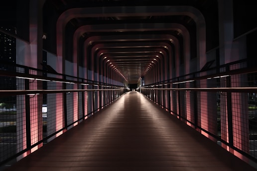 A modern, well-lit pedestrian bridge with a repetitive geometric design, featuring horizontal railings and vertical supports that create a tunnel-like effect. The bridge is illuminated by a series of evenly spaced lights that highlight the structure's symmetrical pattern.