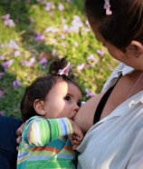 A close-up scene of a breastfeeding moment where a child, wearing a colorful striped shirt, is nursing. The child has a small bow in their hair, and the focus is on the bond between the child and the caregiver. They are outdoors, as indicated by the grass and scattered pink petals on the ground.