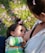 A close-up scene of a breastfeeding moment where a child, wearing a colorful striped shirt, is nursing. The child has a small bow in their hair, and the focus is on the bond between the child and the caregiver. They are outdoors, as indicated by the grass and scattered pink petals on the ground.
