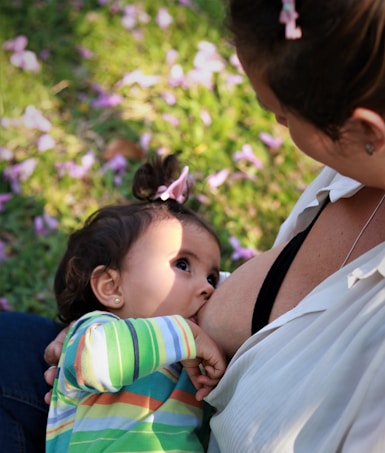 A close-up scene of a breastfeeding moment where a child, wearing a colorful striped shirt, is nursing. The child has a small bow in their hair, and the focus is on the bond between the child and the caregiver. They are outdoors, as indicated by the grass and scattered pink petals on the ground.