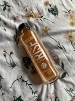 A bottle of RISE Kombucha with orange and turmeric flavor lies sideways on a floral-patterned fabric. The fabric features various flowers in green, yellow, and pink hues.