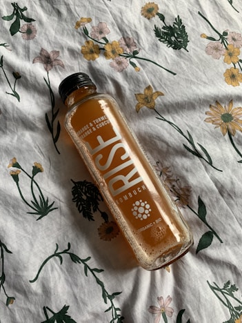 A bottle of RISE Kombucha with orange and turmeric flavor lies sideways on a floral-patterned fabric. The fabric features various flowers in green, yellow, and pink hues.