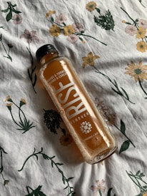 A bottle of RISE Kombucha with orange and turmeric flavor lies sideways on a floral-patterned fabric. The fabric features various flowers in green, yellow, and pink hues.