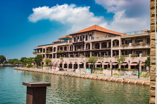 A large Mediterranean-style building with balconies and terracotta roof tiles overlooks a calm waterfront. Trees and plants line the promenade, adding greenery to the scene. The sky is clear and blue, with a few scattered clouds, imparting a serene atmosphere.