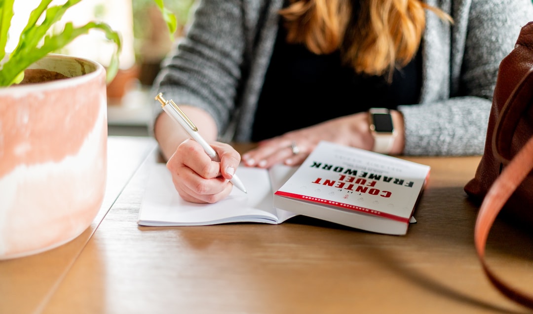 woman in black long sleeve shirt writing on white paper, A woman taking notes on a book at a cafe, with a white pen in her hand, and a plant and a brown purse visible in the edges.