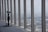 A telescope is positioned near large vertical windows offering a panoramic view of a cityscape with numerous buildings visible in the distance. The interior features a dark carpeted floor, and the windows reflect the overcast sky, lending a subdued atmosphere.