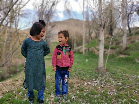 Two young children interact joyfully in a natural setting surrounded by bare trees and patches of grass. The older child wears a teal patterned dress, while the younger child dons a red and green jacket with blue pants. Both children have bright expressions, suggesting a playful conversation.